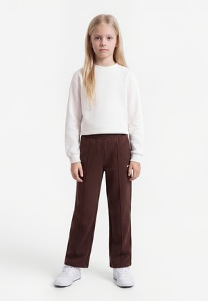 Jeune fille aux longs cheveux blonds debout, portant un sweat-shirt blanc, un pantalon marron et des baskets blanches, sur un fond blanc uni.