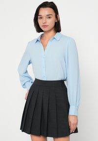 Ανοιχτό γαλάζιο πουκάμισο με κουμπιά και μακριά μανίκια, σε συνδυασμό με σκουρόχρωμη φούστα με πιέτες. Διαθέτει κλασικό γιακά και λεία υφή υφάσματος.