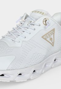 Άσπρο αθλητικό παπούτσι Guess με υφασμάτινη υφή, χρυσό λογότυπο στη πλευρά, μπροστινό δέσιμο με κορδόνια και μοναδικό σχεδιασμό αεριζόμενης σόλας.