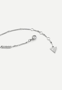 Bracciale in argento con logo "GUESS" e un ciondolo triangolare. La catena presenta maglie con piccoli accenti circolari e un moschettone.