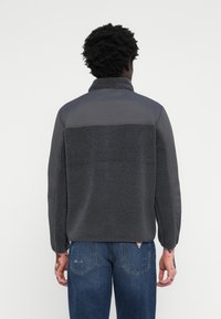 Veste en polaire grise avec un haut matelassé gris. Elle présente un col haut et une coupe décontractée. Portée avec un jean bleu dégradé.