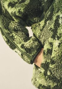 Zelena krznena jakna koja se ističe teksturiranim, čupavim materijalom u različitim nijansama zelene. Sadrži džep za ruke i završene rubove.