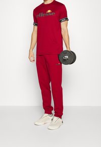 Rotes Sportoutfit bestehend aus einem kurzärmligen T-Shirt mit Logo, passenden Hosen und weißen Sneakers. Hält ein schwarzes Gewicht.