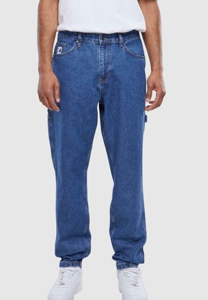 Vaqueros de mezclilla azules con un corte relajado, que cuentan con bolsillos laterales, un parche de logo en el frente y puños elásticos, estilizados con zapatillas blancas.