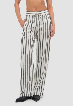 Frau trägt weiße Hose mit schwarzen vertikalen Streifen, elastischem Bund mit Kordelzug und schwarzen spitzen Schuhen, steht mit einer Hand in der Tasche.