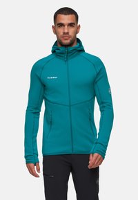 Giacca in pile zip-up color teal con colletto alto, design fitted e logo sul petto. Presenta aperture per i pollici e due tasche laterali con zip.