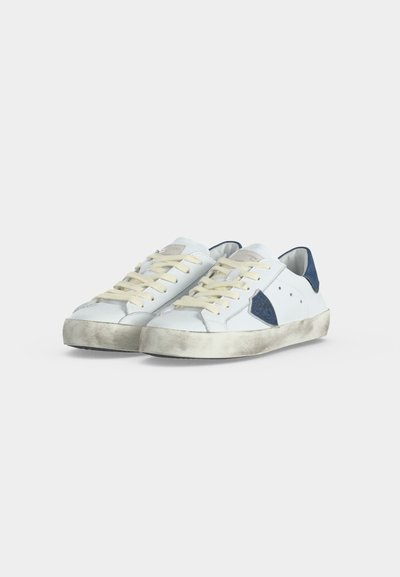 Paire de baskets basses blanches avec des lacets écrus, des semelles abîmées et des détails bleus au talon, placées côte à côte sur un fond uni.