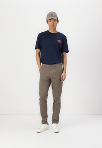 Marineblaues T-Shirt mit orangefarbenem Grafikdesign, khakifarbene Chinos und weiße Sneakers. Die Accessoires umfassen eine graue Baseballmütze. Das Model steht vor einem schlichten weißen Hintergrund.