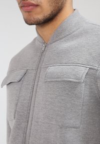 Close-up of a man wearing a light grey zip-up jacket with two flap chest pockets and a ribbed collar.