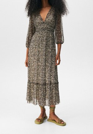 Femme portant une longue robe à motifs floraux avec des manches trois-quarts en voile transparent et des sandales plates à lacets vert olive.