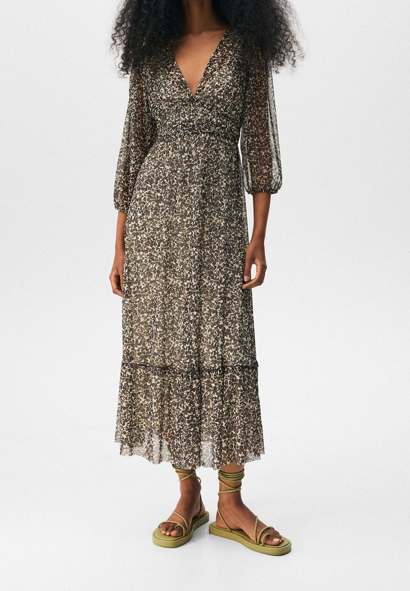 Femme portant une longue robe à motifs floraux avec des manches trois-quarts en voile transparent et des sandales plates à lacets vert olive.