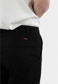 Schwarze Hose mit glatter Textur, versehen mit einem kleinen, orangefarbenen gestickten Detail. Taschen sind an den Seiten und hinten sichtbar.