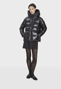 Svart vadderad jacka med glansigt ytskikt, huva och dragkedja framtill, med quiltade paneler. Bärs över en svart kjol och strumpbyxor.