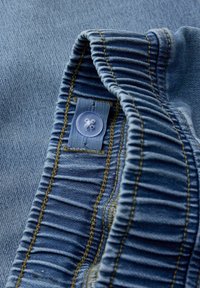 Jeans tkanina svetlo modre barve, z nabrano teksturo, plastičnim gumbom in zlatim šivom ob robovih.