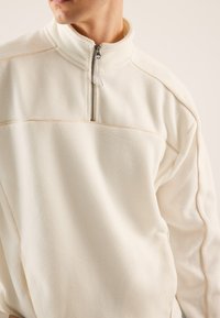 Balts polārfleeces džemperis ar augstu apkakli un pusautomātisku rāvējslēdzēju, ar kontrastējošām bēšām šuvēm gar šuvēm. Mīksta tekstūra.