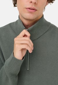 Gerippter grüner Reißverschluss-Pullover mit hohem Kragen. Verfügt über einen Metallreißverschluss und eng anliegende Bündchen, die eine weiche Textur zeigen.