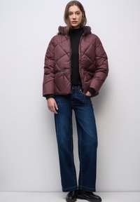 Burgunderfarbene Steppjacke mit einer taillierten Kapuze, Reißverschlusstaschen und rautenförmigem Steppmuster. Getragen über einem schwarzen Rollkragenpullover und blauen Jeans.