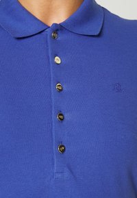 Polo azul hecho de tejido de punto, con cuello clásico, cinco botones dorados en la tapeta y un logo bordado en el pecho.