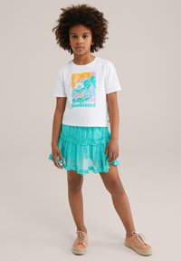 Biele tričko s krátkym rukávom a grafikou "Sunkissed"; viacvrstvová tyrkysová sieťová sukňa s volánmi a kvetinovým vzorom; béžové šnurovacie topánky po členky.