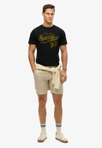 Sort t-shirt med gyldent trykt logo, beige shorts, cremefarvet sweater bundet om livet, og lyse sneakers med detaljer.