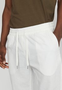 Hombre vestido con pantalones de lino blancos con cordón y camiseta verde oliva, con la mano derecha en el bolsillo y la mano izquierda relajada a un lado.