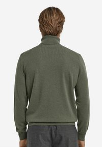 Dunkelgrüner Pullover mit Stehkragen, strukturiertem Strickmaterial, gerippten Ärmelbündchen und Saum. Trägt graue Hose, von hinten betrachtet.