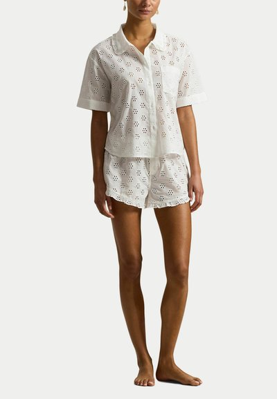 Vrouw die een wit kortermig overhemd met knopen en bijpassende shorts draagt met een gebloemd opengewerkt patroon, blootsvoets, staand tegen een effen achtergrond.