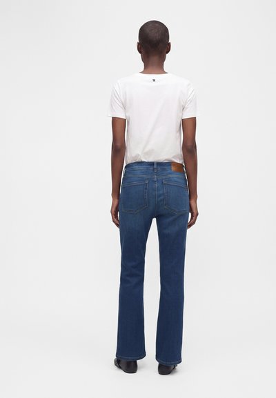 Personne vue de dos portant une chemise blanche à manches courtes, un jean bleu avec des poches arrière, et des chaussures noires, debout sur un fond clair uni.