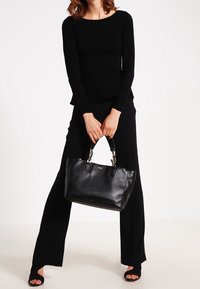 Sac fourre-tout en cuir noir au design structuré, avec une quincaillerie argentée et des doubles poignées, tenu par une personne portant un haut noir et un pantalon à jambes larges.