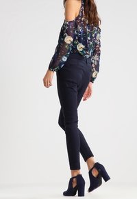 Genomskinlig marinblå blus med blommönster och bara axlar, kombinerad med figurnära svarta byxor och mörkblå klackar, som visar en slank silhuett.