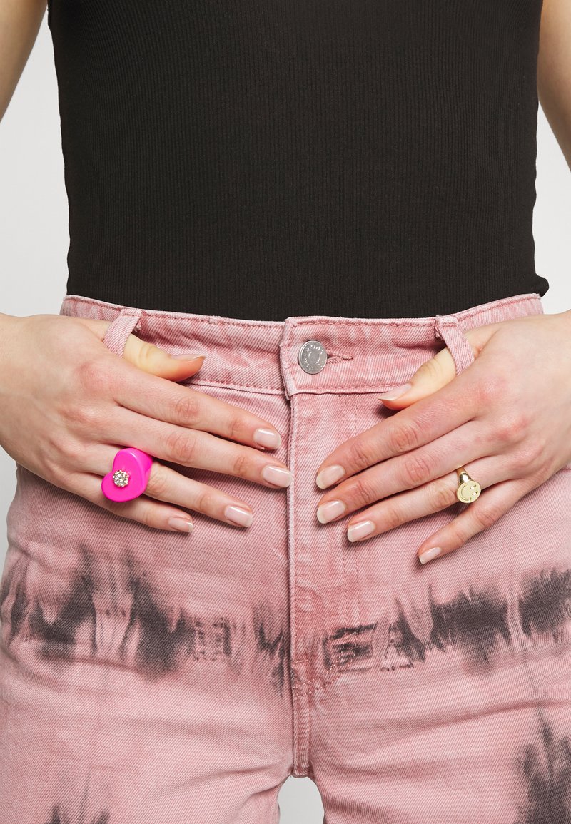 Calças de ganga cor-de-rosa com um padrão de tie-dye, top preto com textura canelada, grande anel cor-de-rosa com gema, e um anel dourado com um design de carinha sorridente nas mãos.