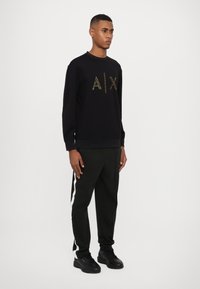 Sudadera negra con logo dorado "AIX", combinada con pantalones verde oscuro que tienen rayas laterales blancas y zapatillas negras. Diseño simple y deportivo.