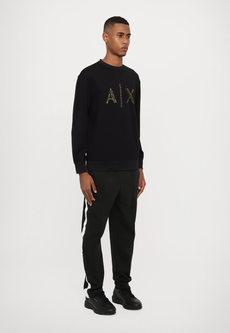 Schwarzer Sweatshirt mit goldenen "AIX"-Logo, kombiniert mit dunkelgrünen Hosen, die weiße Seitestreifen haben, und schwarzen Sneakers. Einfaches, sportliches Design.