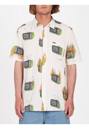 Hombre vistiendo una camisa blanca de manga corta con botones y estampado de televisores retro verdes en llamas, combinada con jeans de mezclilla azul claro.