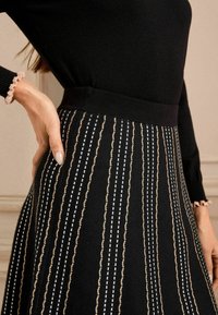 Falda negra con rayas verticales en blanco y marrón claro. Presenta una cinturilla ajustada y un tejido texturizado. Primer plano de una mano al lado.