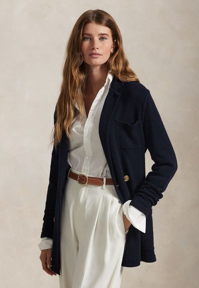 Donna con capelli castani lunghi indossa una camicia bianca e pantaloni con cintura marrone e un cardigan blu navy su uno sfondo beige uniforme.