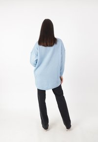 Suéter de punto de gran tamaño en azul claro con un dobladillo redondo, usado sobre pantalones negros. Presenta un tejido texturizado y un diseño de hombro caído.