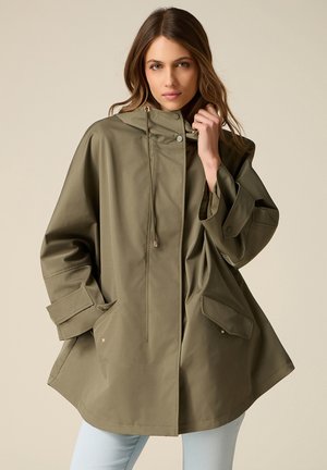 Femme portant un imperméable ample à capuche vert olive avec de grandes poches et des poignets ajustables, posant devant un fond neutre.