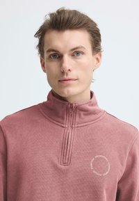 Lichtroze zip-up sweater met een hoge kraag, voorzien van een ronde witte tekstlogo op de borst en een zachte, gestructureerde stof.