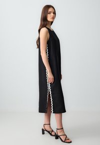 Zwarte mouwloze jurk met een rechte silhouet, voorzien van een decoratieve witte zigzag trim aan de zijkant en een hoge split. Gecombineerd met zwarte hoge hakken sandaaltjes.