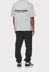 Šedé bavlněné tričko s černým nápisem "PEGADOR" na zádech, spárované s černými tepláky a tmavě modrými teniskami Adidas s bílými akcenty.