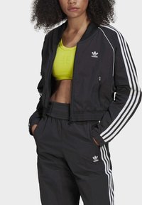 Giacca bomber nera con strisce bianche sulle spalle, tasche con zipper e polsini elasticizzati. Abbinata a un crop top giallo brillante e pantaloni neri abbinati.