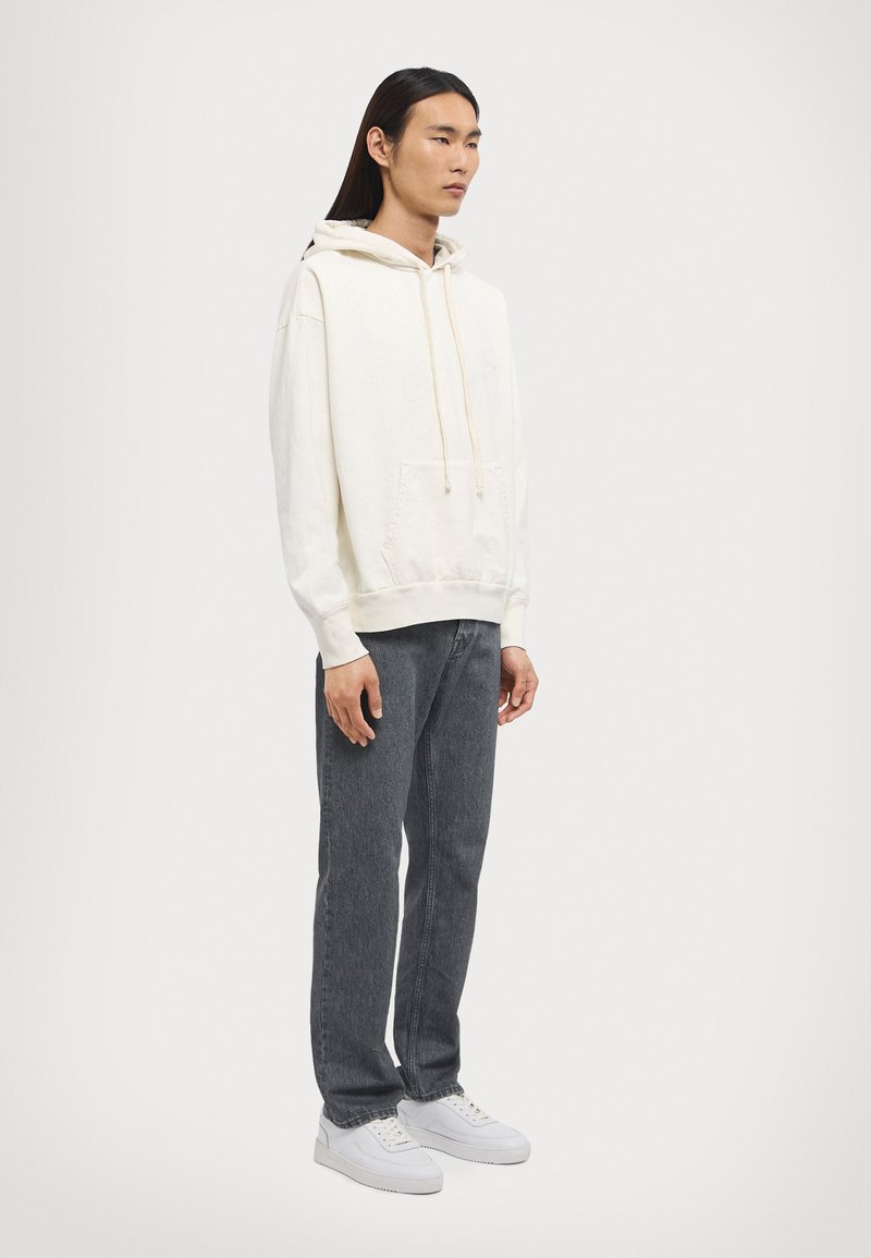 Hooded sweatshirt in cream with a kangaroo pocket, paired with dark gray jeans and white sneakers. Simple design, soft texture.