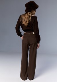 Jeans marroni a gamba larga con vita alta e tasche posteriori, abbinati a un maglione scuro e a un cappello a secchiello in pelliccia. Tessuto dalla trama liscia.