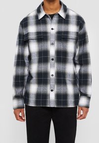 Camicia di flanella a quadri neri, bianchi e grigi con chiusura con bottoni, due tasche sul petto e colletto classico. Il tessuto sembra morbido con una leggera texture.