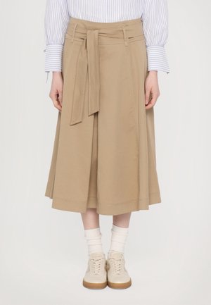 Kvinde iført beige A-linje midi-nederdel med stofbælte, kombineret med hvide sokker, beige sneakers og en blåstribet langærmet skjorte.