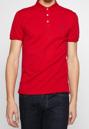 Mann trägt ein eng anliegendes rotes Poloshirt mit kurzen Ärmeln, drei weißen Knöpfen und dunkelblauen Jeans vor einem einfarbigen Hintergrund.