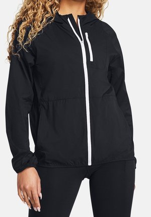 Kvinde iført en sort lynlåsjakke med hvide lynlåse og elastiske ærmegab, kombineret med sorte leggings.