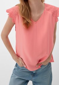 Blouse sans manches corail avec bordure en dentelle festonnée sur les épaules, encolure en V et tissu doux et léger. Portée avec un jean décontracté.