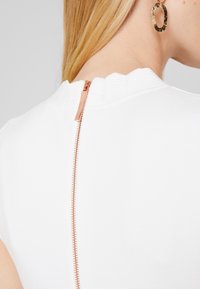 Achteraanzicht van een persoon die een wit topje draagt met een roségouden rits en een geschulpte halslijn, en een gouden ovale oorbel.